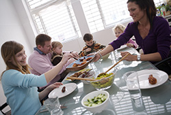Mange barn og to voksne spiser mat ved et stort bord. Foto.