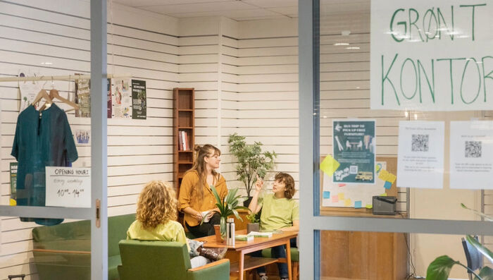 En åpen glassdør med plakat som sier Grønt Kontor. På innsiden av døren sitter tre studenter i en grønn sittegruppe og prater sammen.