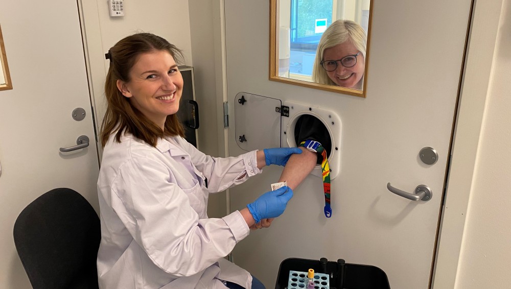 Image shows a woman in a lab coat taking a blood samle from a woman behind a door, putting her arm through a hatch.