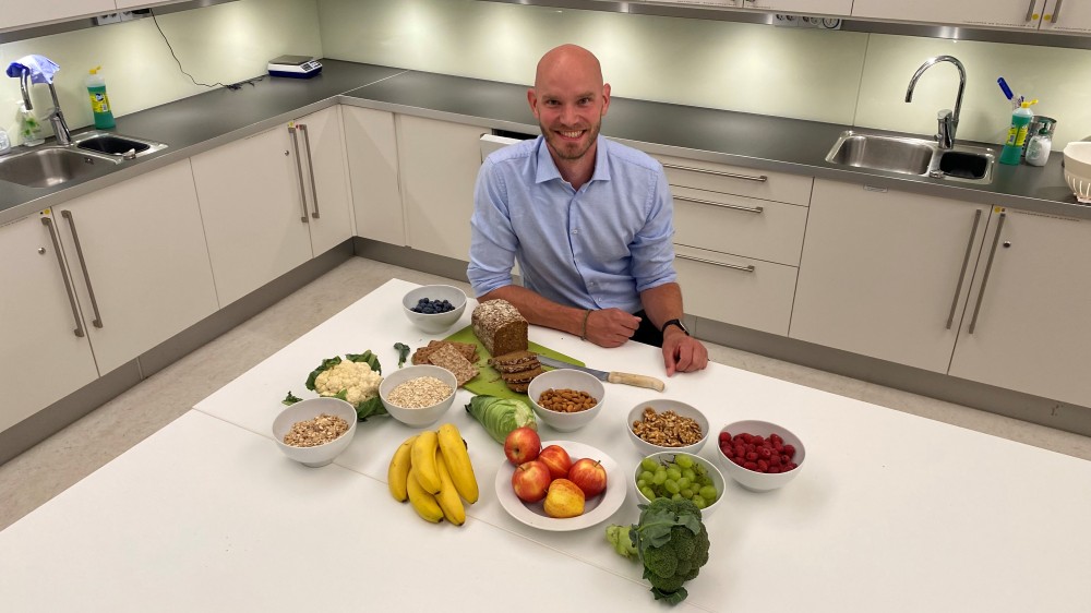 Image shows a man sitting at a table filled with vegetables, fruit and bread