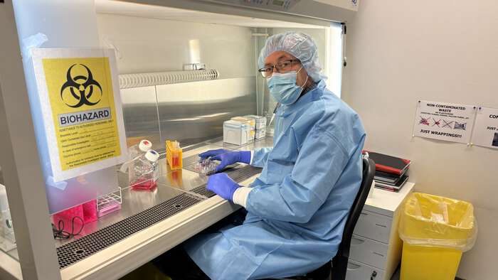 Image shows a man in blue lab coat, face mask and gloves working in a lab