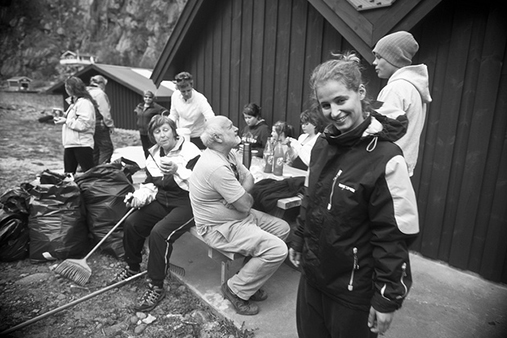 Eleven people outside gathered around a bench with black plastic bags and rakes. Photo.