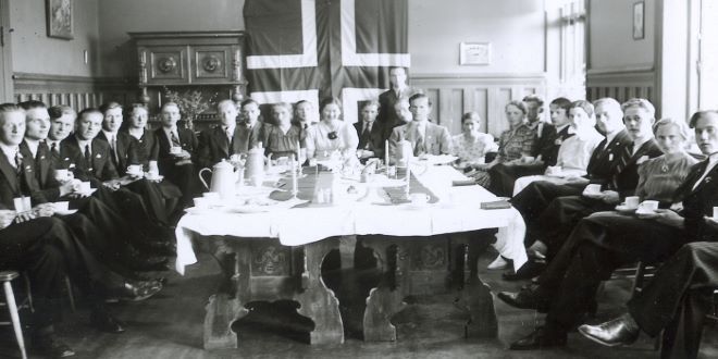 Black and white photo. Men and women are gathered around a big table. The Norwegian flag is hanging on the back wall.