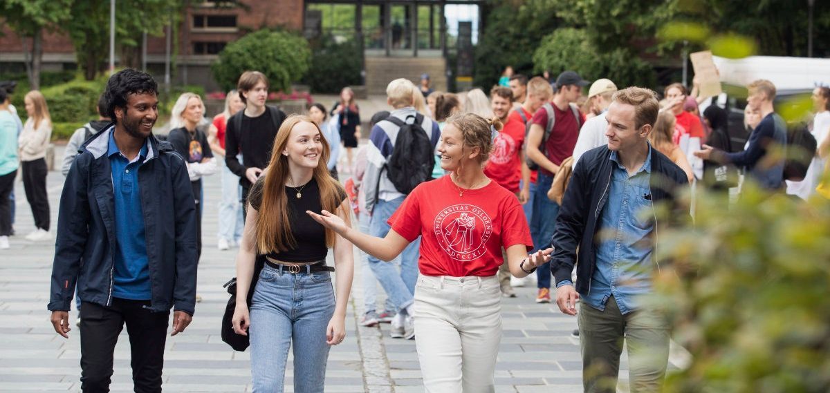 UiO studenter går side om side mot fotografen. I midten en fadder i UiO t-skjorte som slår hendene ut til sidene og snakker med de andre.