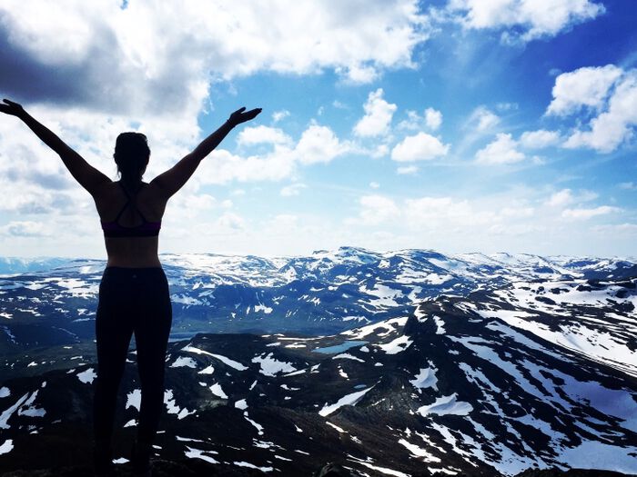 person standing on mountain with arms above head looking out over mountains with blue sky and fluffy white clouds above