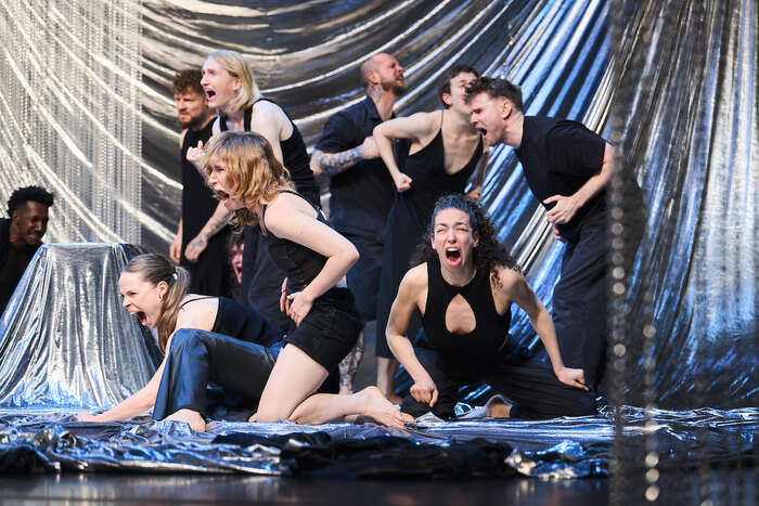 En gruppe mennesker i sorte klær utfører en energisk danseopptreden på en scene med glitternde gardiner.