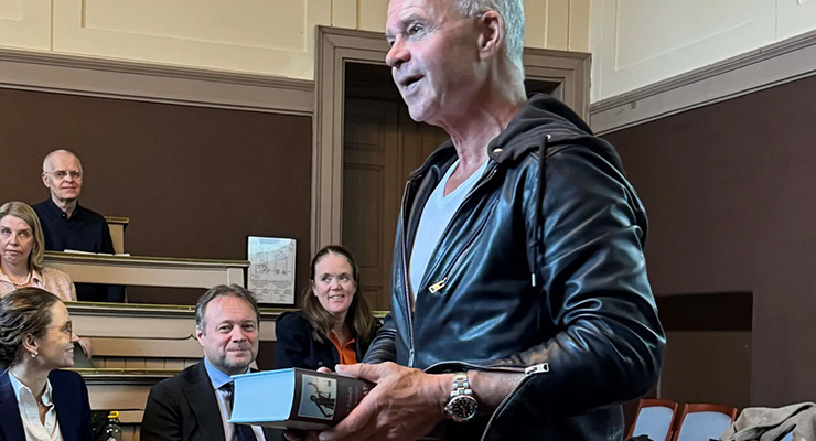 Mann i svart skinnjakke holder tykk bok i auditorium.