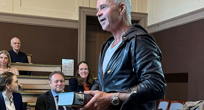 Mann i svart skinnjakke holder tykk bok i auditorium.