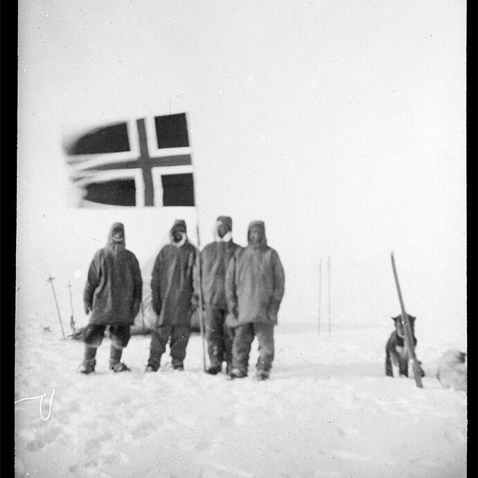 Fire godt kledde menn står i isødet på Sydpolen ved et norsk flagg som står plantet i snøen. Ved siden av dem står en hund. 