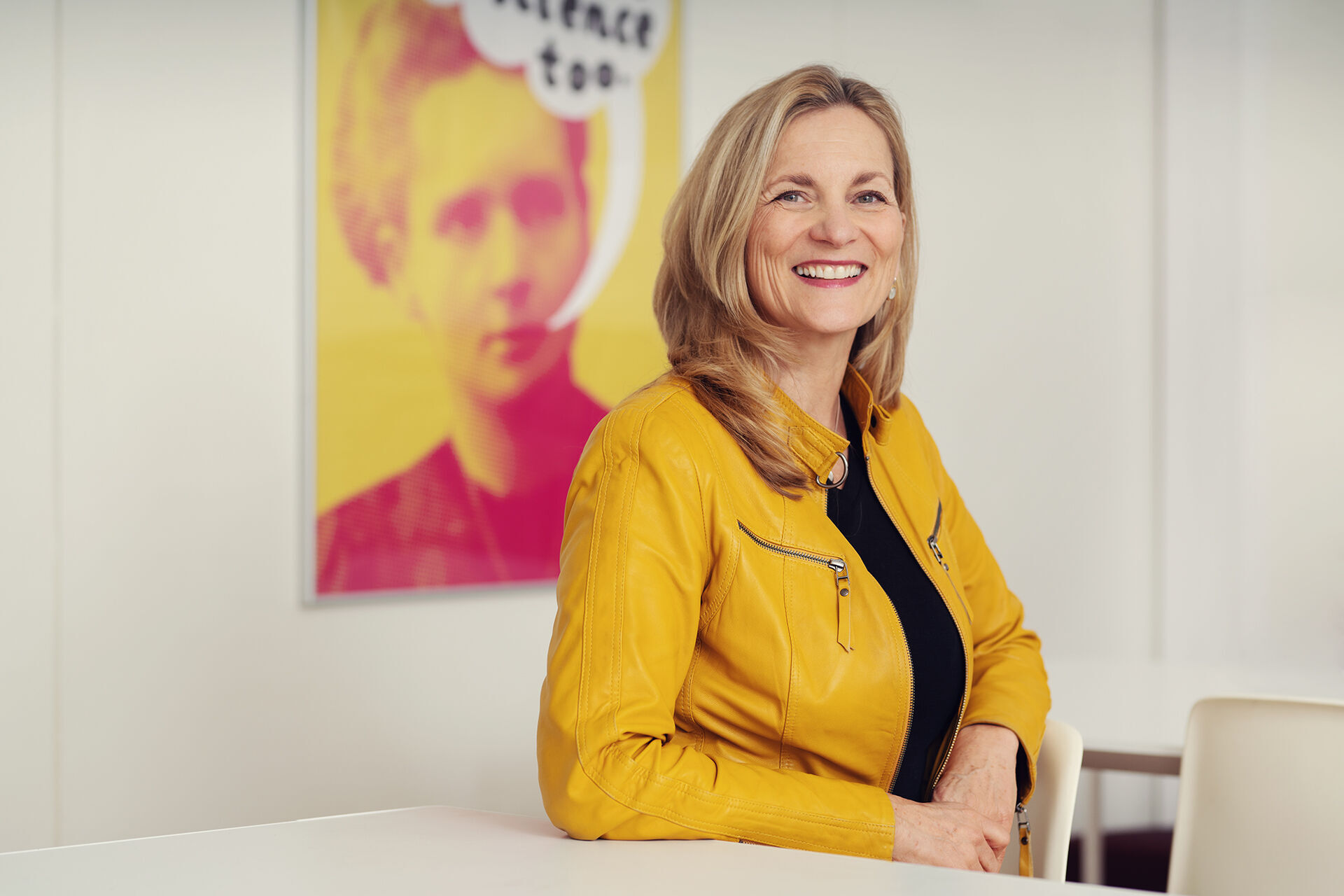 En smilende dame med lyst hår, gul jakke og sort genser som sitter ved et bord