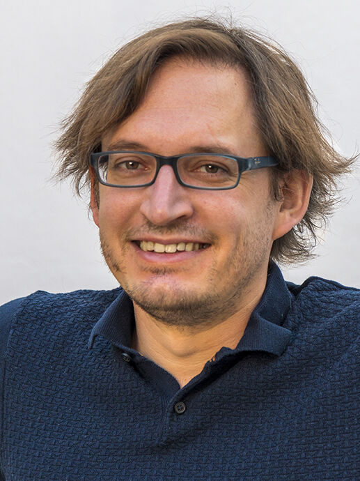 Portrait of a man with medium blond hair and glasses looking at the camera and smiling gently. Photo.