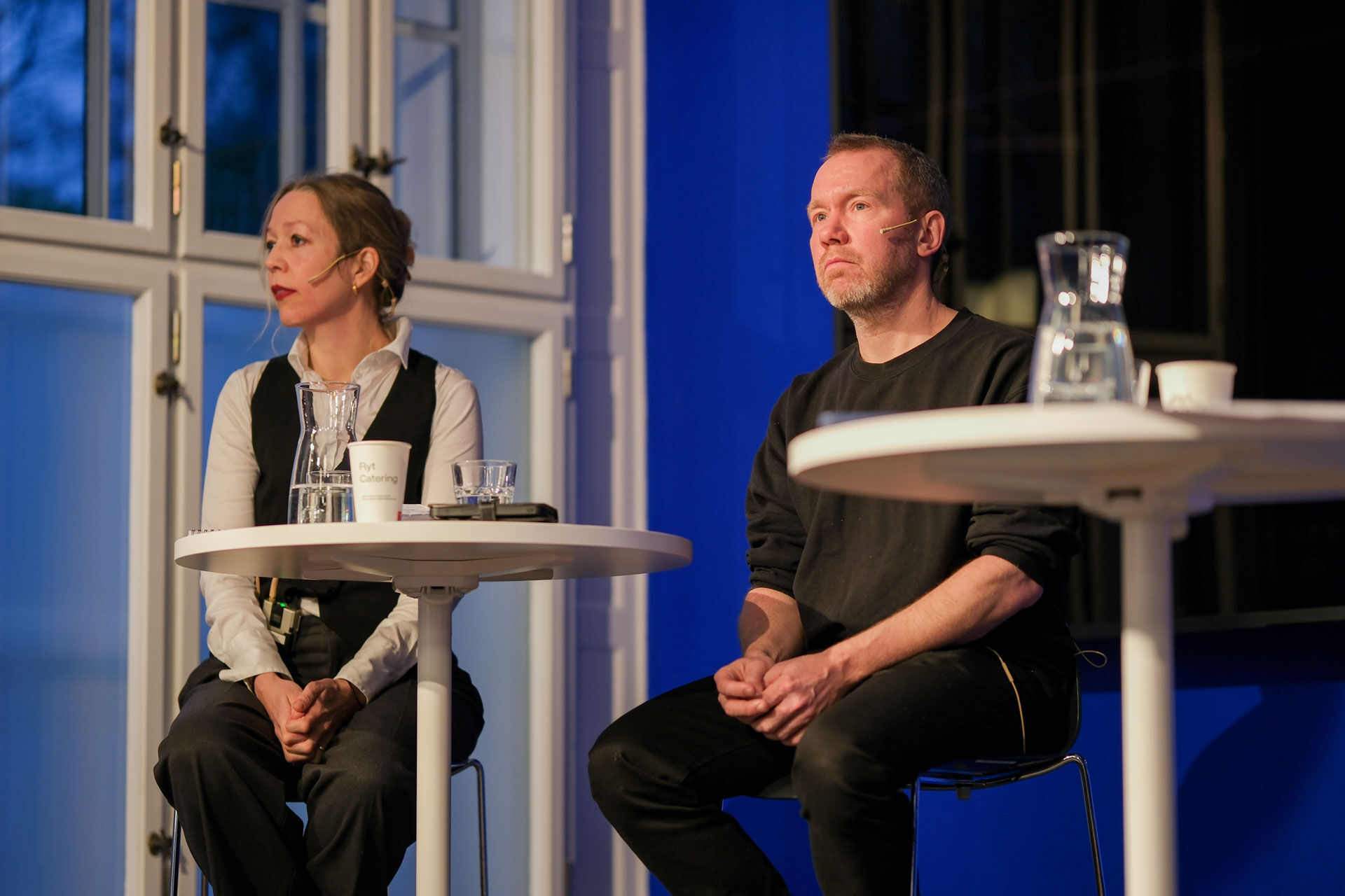 Kristin Bertora Sandvik og Jonas Bals sitter på to høye stoler i et blått rom og snakker til publikum.