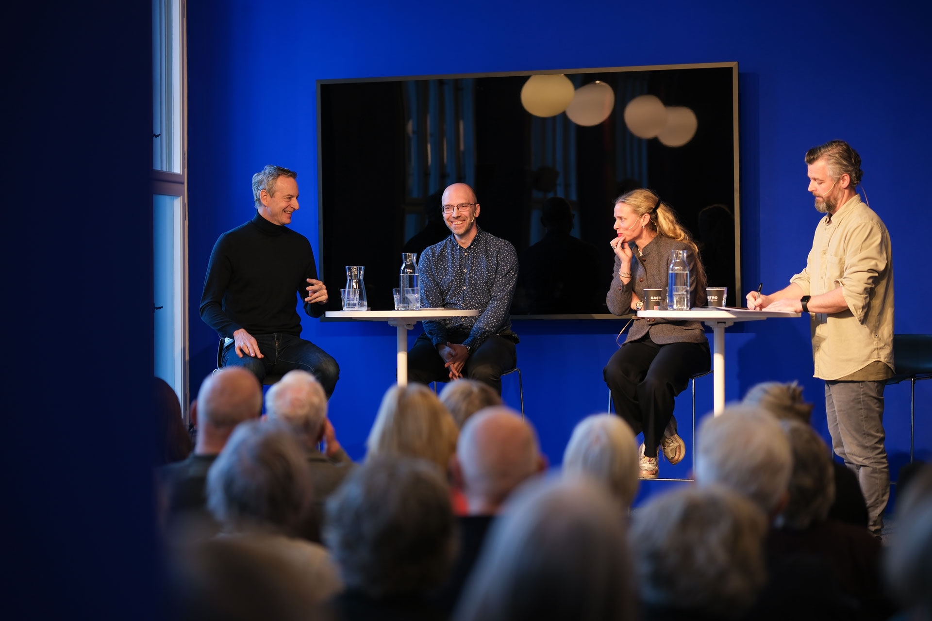 Karsten Friis, Jørgen Jensehaugen. Marte Heian-Engdal og Arild Blomkvist står på scenen i et blått rom. Fulle rader med publikum sitter og hører på. 