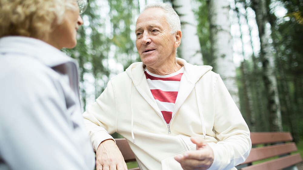 To eldre personer sitter på en benk i skogen. Den ene har kort, grått hår, lys hettegenser og en rød‑ og hvitstripet genser, og vender seg mot den andre mens han snakker. Foto.