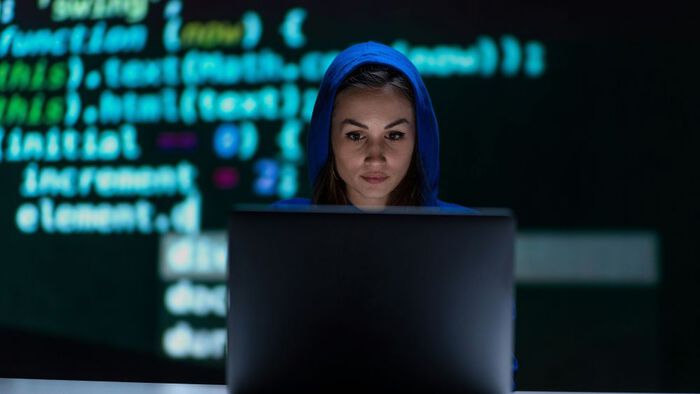 En kvinne med hettegenser forran en pc-skjerm. I bagrunnen digital skrift på en stor skjerm.