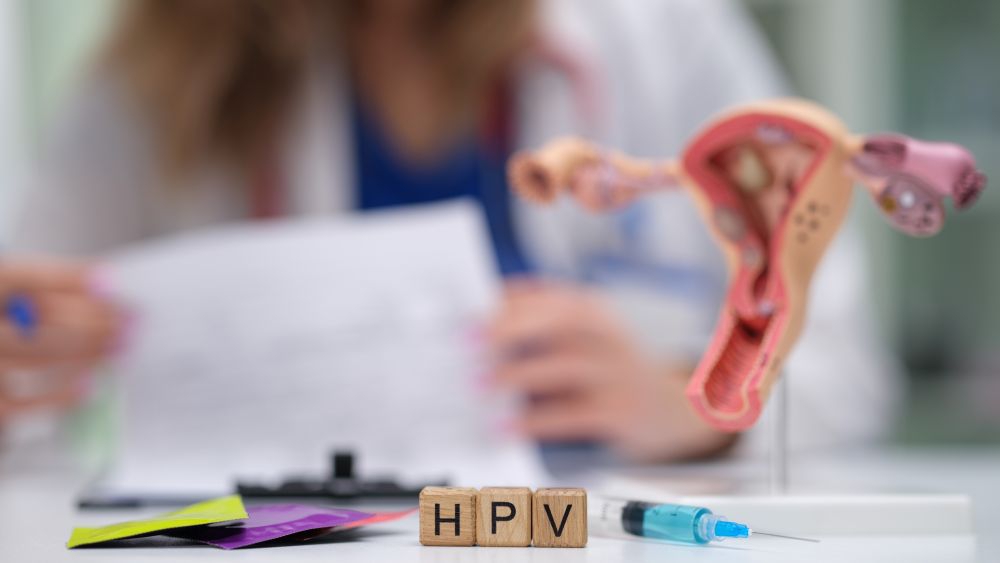 Lege leser på et papir, foran seg en modell av livmorhalsen og tre terninger med bokstavene HPV