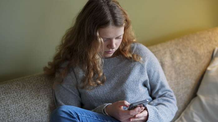 Jente med langt hår  som titter ned i mobilen sin. Sitte rpå en sofa 