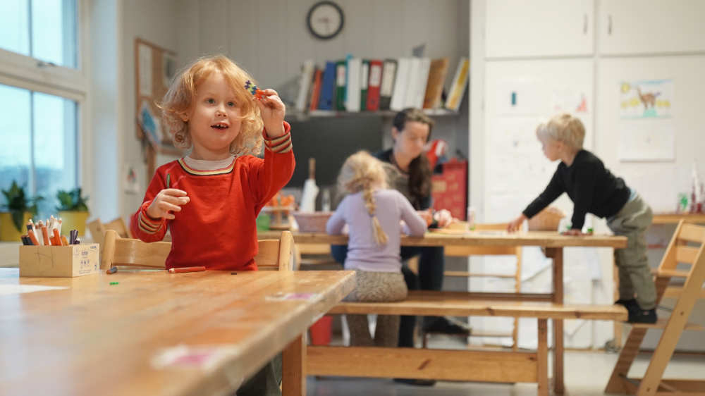 To barnehagebarn sitter ved et bord og leker sammen med en barnehageansatt, mens et annet barn leker alene ved et annet bord.