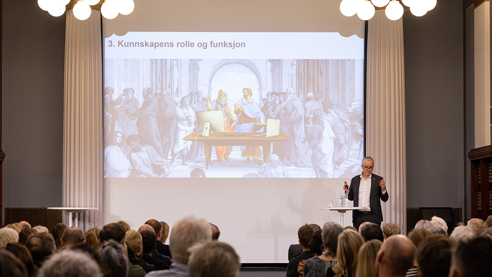 En mann står på scenen og snakker til fullsatt sal.