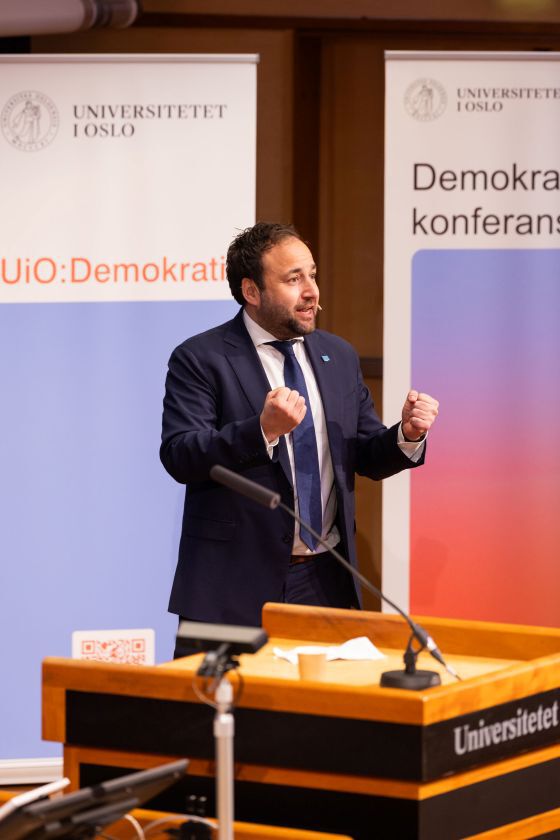 Mann med dress står med knyttede never og snakker. Bak er to bannere med UiO-logo og teksten UiO:Demokrati. Foto. 