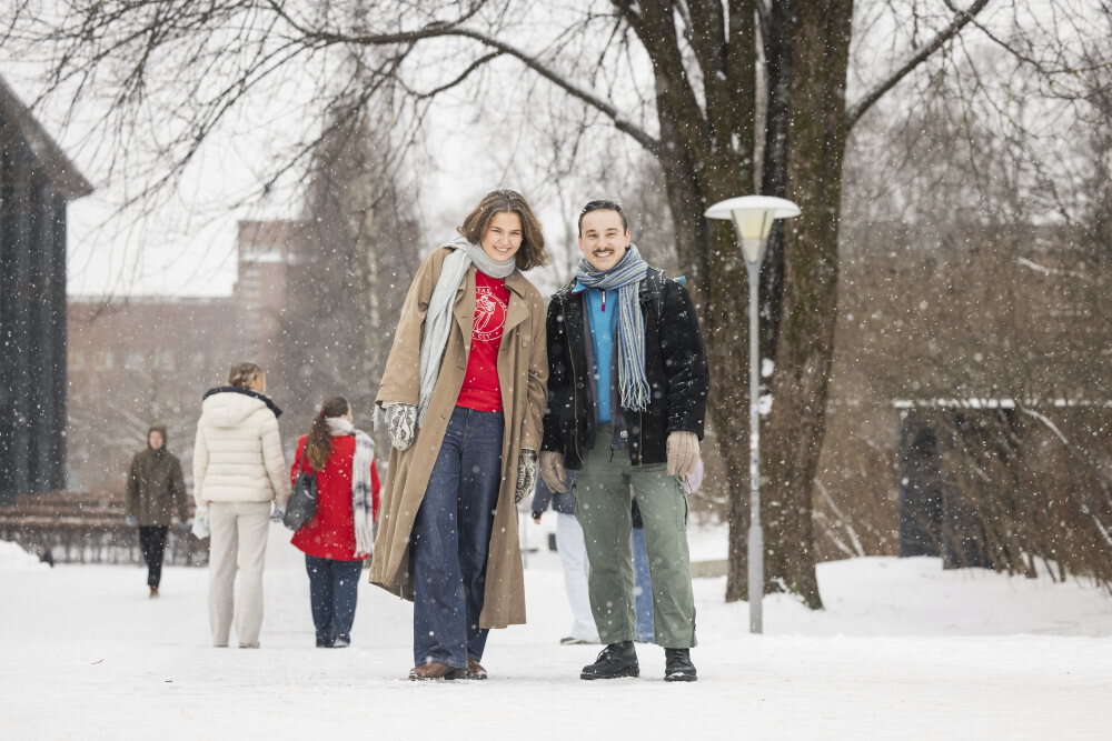 To studenter på campus. De har på seg vinterklær, og en av dem en fadderskjorte. Det snør. 