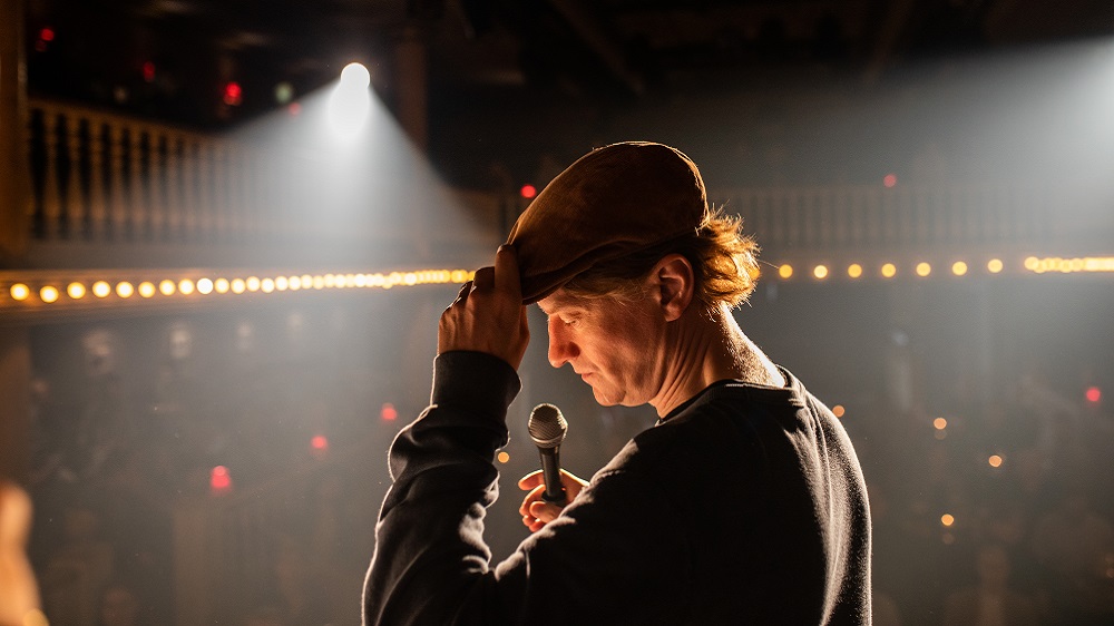 En mannlig standupkomiker med sixpence står på en scene foran et publikum. Han har mikrofon i hånden. Foto.