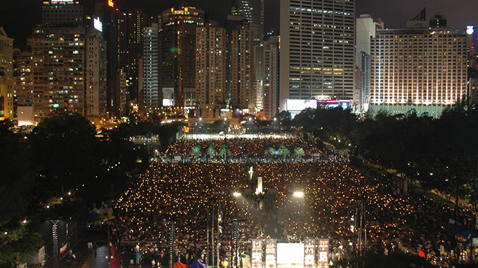 Avstandsbilde på kveldstid av store folkemengder med lys i en park omgitt av skyskrapere.