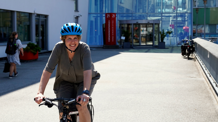 Kvinne med blå sykkelhjelm på sykkel, sterk sommersol. Hun smiler,
