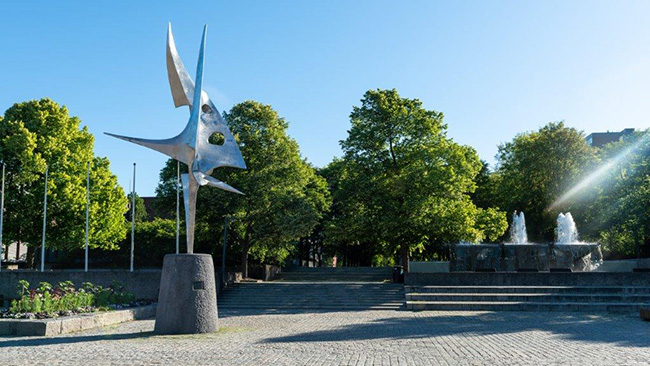 Stor skulptur på en plass med grønne trær i bakgrunnen