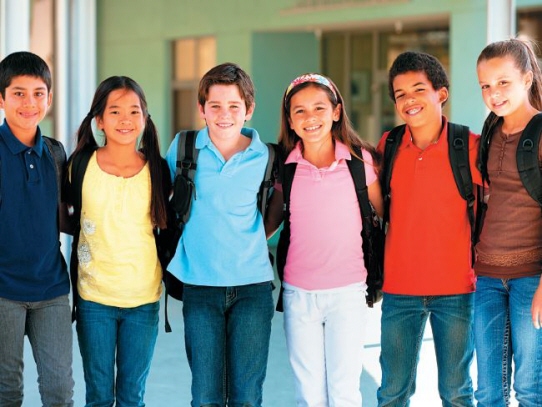 Seks oppstilte barneskoleelever i fargesterke trøyer med skoleransler på ryggen. Foto.