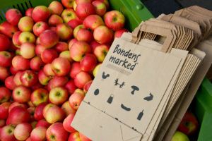 Nærbilde av en kasse med rød-gule epler og gråpapirposer med påskriften "Bondens marked".