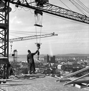 En mann på toppen av en byggeplass som holder i en krok som er festet i en heisekran. Sort-hvitt-foto.