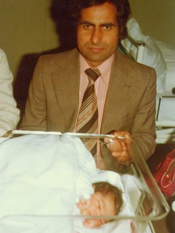 En liten baby i en sykehustralle sammen med en mann med mørkt hår og gul-brun dress. Foto.