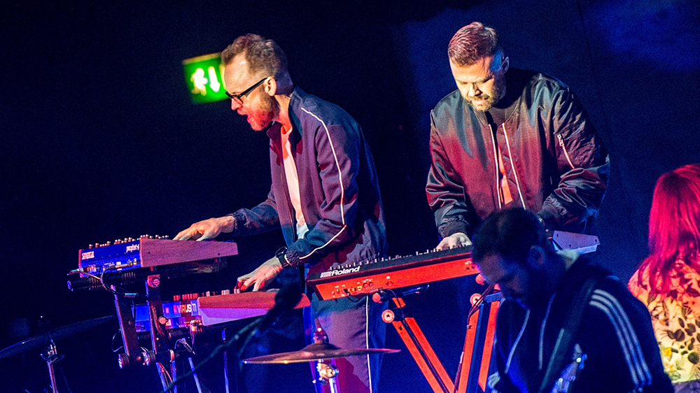 Two men are standing on a stage behind their respective keyboards. They are playing. In the foreground, there is a man playing bass. Photo.