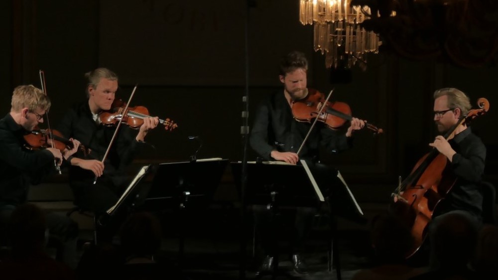 The four members of The Danish String Quartet playing at "Musikhuset" (The Music House) in Copenhagen. The focus is on the quartet, with chandeliers in the foreground to the right and in the background towards the middle. The room is dimly lit. You can also see the back of the heads of some of the audience.