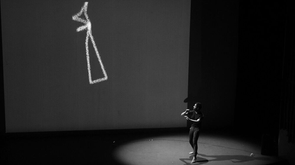 Black and white photo of Diego dancing on a stage with a projection screen behind him showing an AI avatar in the form of white lines