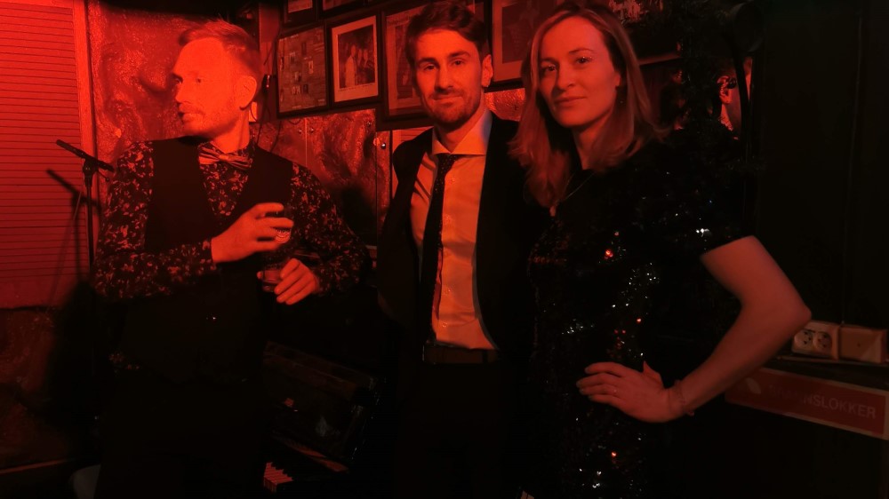 A woman to the far right, two men to the left of her. The men are dressed in suits and the woman dressed in a sequin dress. The room is dimly lit with a red light.