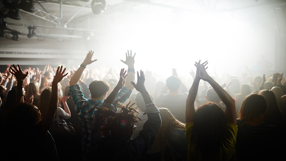 Many people facing a stage with their hands up at a concert. The stage is obscured with a blinding light.