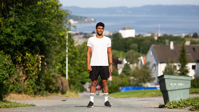 Dennis sittende i hvit t-skjorte med denimbukser. Programlederen i Newton har studert Biologi ved UiO