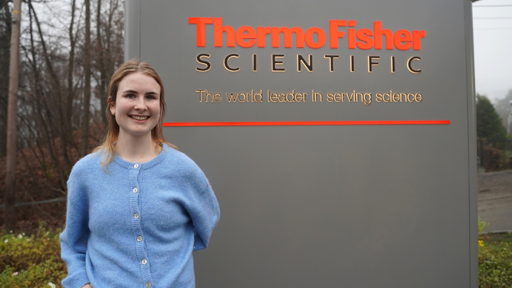 Sunniva står foran skiltet til Thermo Fisher. Smiler. Blå genser. 