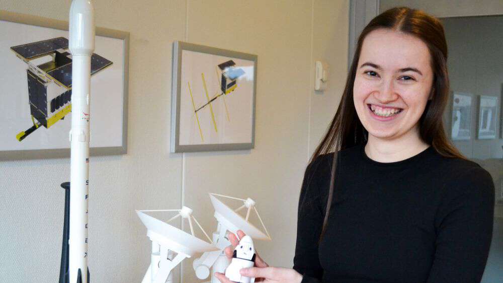 Ung dame med brunt hår smiler til kamera mens hun holder en liten modell av en radarsatelitt på kontoret til arbeidsplassen Space. 