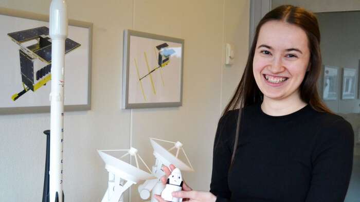 Ung dame med brunt hår smiler til kamera mens hun holder en liten modell av en radarsatelitt på kontoret til arbeidsplassen Space. 