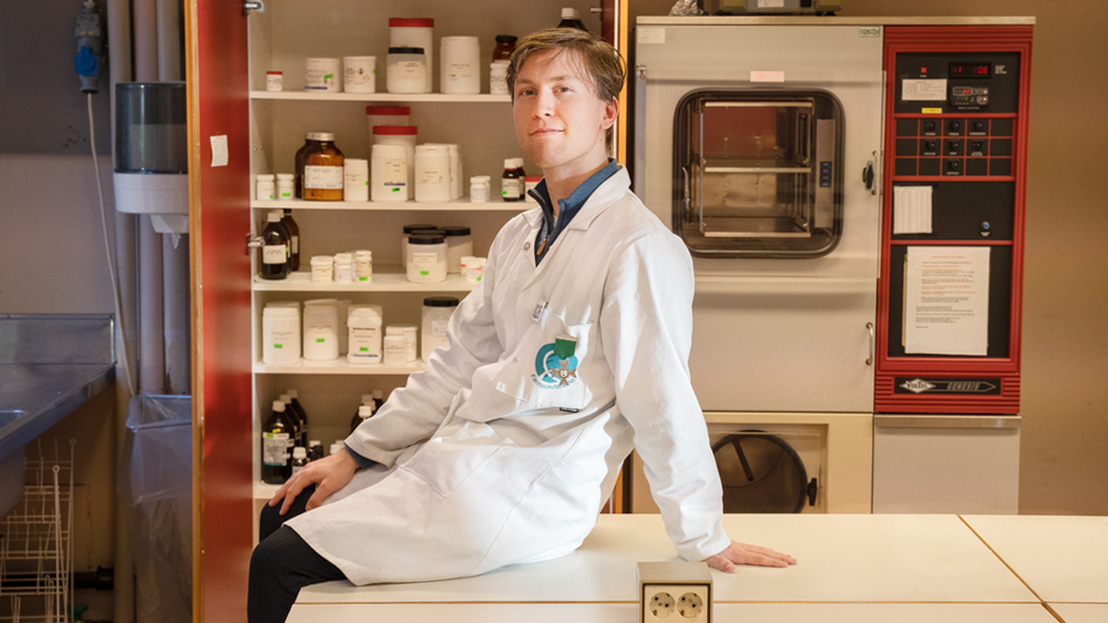 Blond mann som sitter på et bord framfor et skap med medisiner i. 