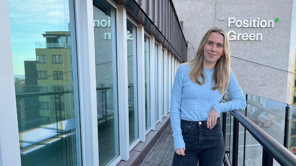 Jente i blå genser og blondt hår står på en takterasse og smiler til kameraet. 