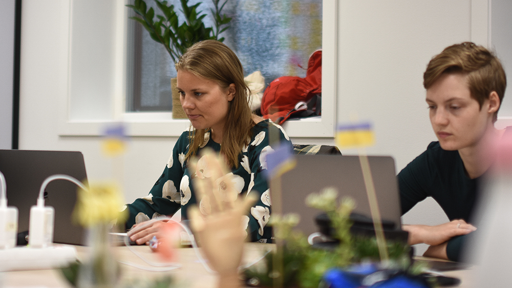To kvinnelige studenter sitter og jobber i et lyst kontorlandskap med blomster og ledninger