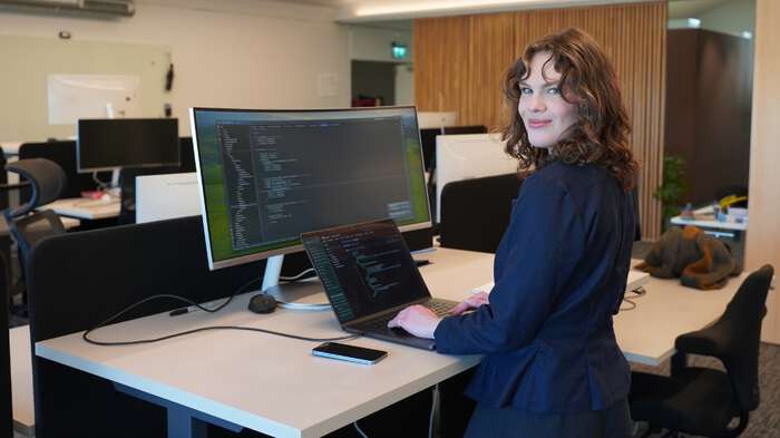 Ung dame med krøllete hår og mørkeblå topp smiler til kamera mens hun står foran en pc-skjerm. 