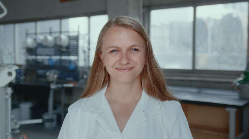 En ung kvinne med blondt hår og hvit laboratoriefrakk smiler mot kameraet.