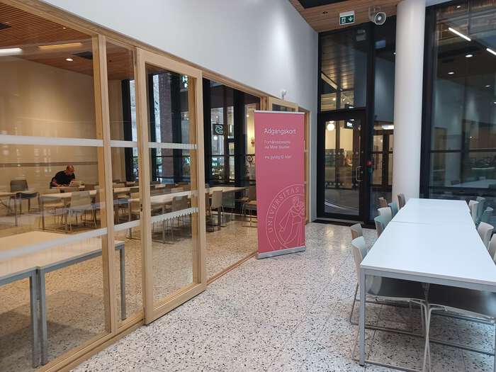 Bildet viser glassdører inn mot et rom hvor det sitter en mann ved et bord, på en rød banner ved døren står det Adgangskort.