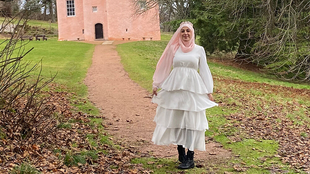 Bilde av student Miriam i hvit kjole og rosa hijab foran et rosa slott i Skottland.