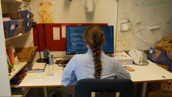Ung dame med brun, lang flette og lyseblå skjorte sitter ryggen til ser på en pc-skjerm.